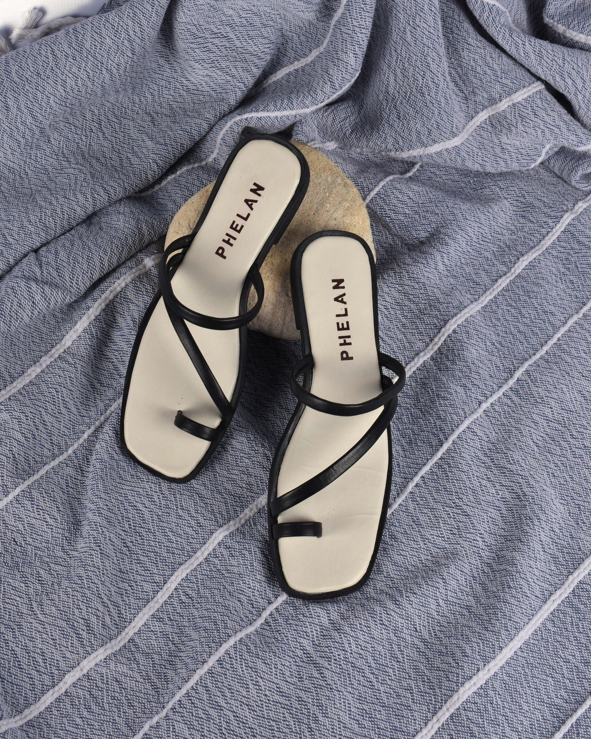 Lipari Slide in Black, the pair on a blue-grey throw with a sandstone pebble